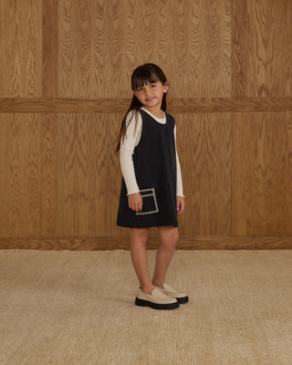 Rylee & Cru Ribbed Long Sleeve - Ivory
Young girl wearing a black dress with a white shirt underneath, standing against a wooden paneled wall.