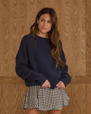 Rylee & Cru Womens Carolina Sweater - Navy Woman wearing a navy sweater and checkered skirt against a wooden paneled wall