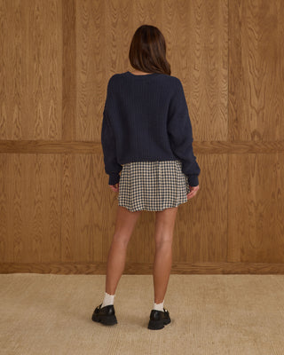 Rylee & Cru Womens Pleated Mini Skirt - Navy Check Person wearing a navy sweater and checkered skirt against a wooden paneled wall.
