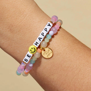 Little Words Project Bracelet - Be Happy
A close-up of a person's wrist wearing three beaded bracelets. One bracelet has white letter beads spelling 'HAPPY', a yellow smiley face charm, and gold spacer beads.