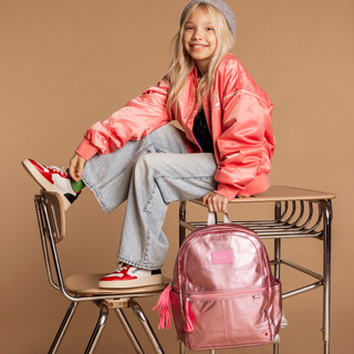 State Bags Kane Backpack - Pink / Silver Young girl in pink jacket and jeans sitting on a school desk with a pink backpack, against a brown background