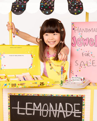Super Smalls Super Entrepreneur Bead Kit Child behind a lemonade stand with jewelry display, wearing a colorful apron.
