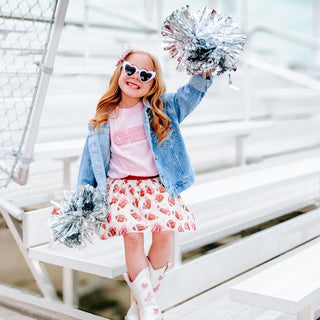 Sweet Wink Game Day Football Sequin Patch Denim Jacket Young girl in a playful pose with pom-poms on a white swing set
