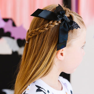 Sweet Wink Glitter Bat Halloween Coquette Bow Clip
Child with braided hair wearing a large black bow against a colorful background